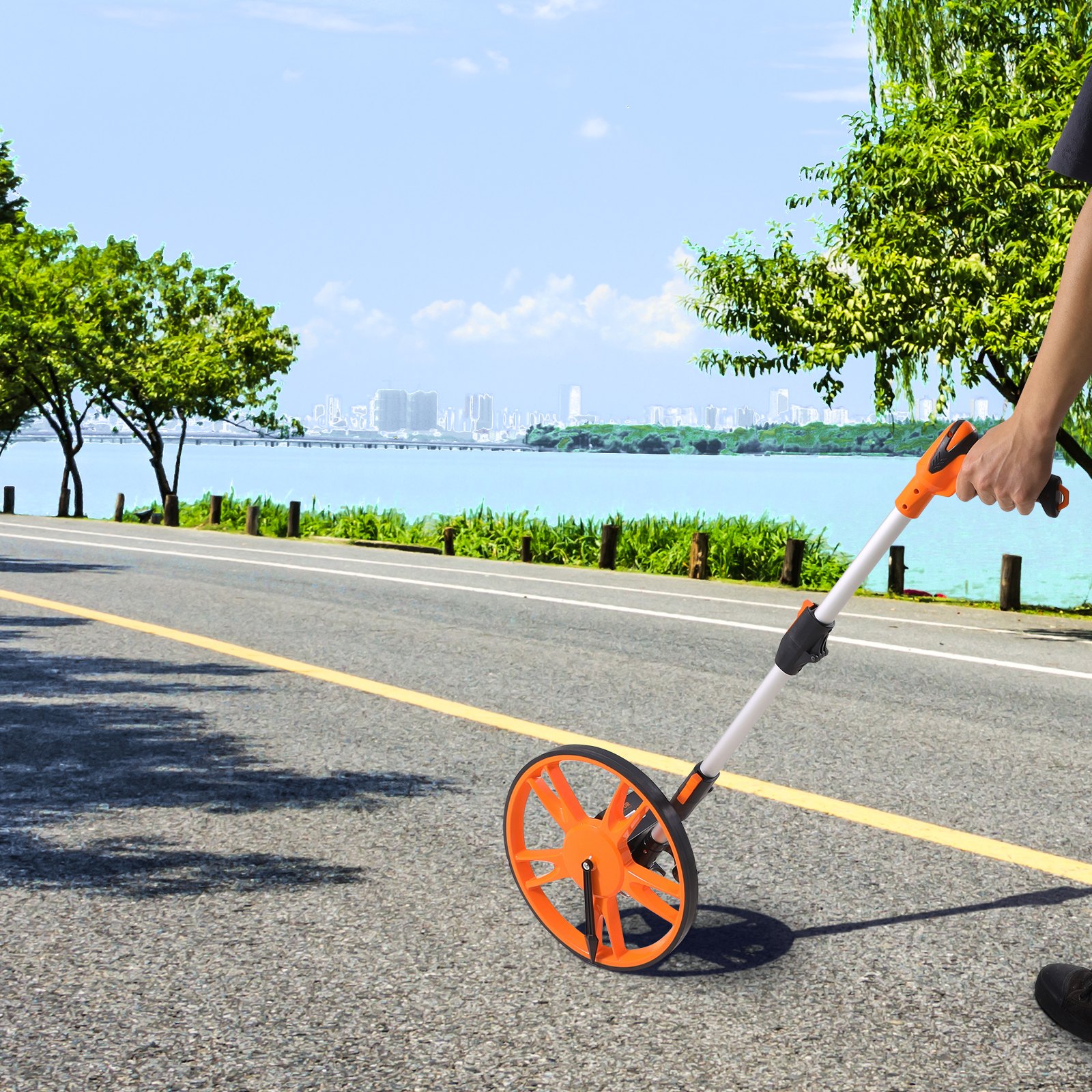 Persona que utiliza la rueda de medición VEVOR en una carretera costera con un horizonte de ciudad y frondosos árboles en el fondo.