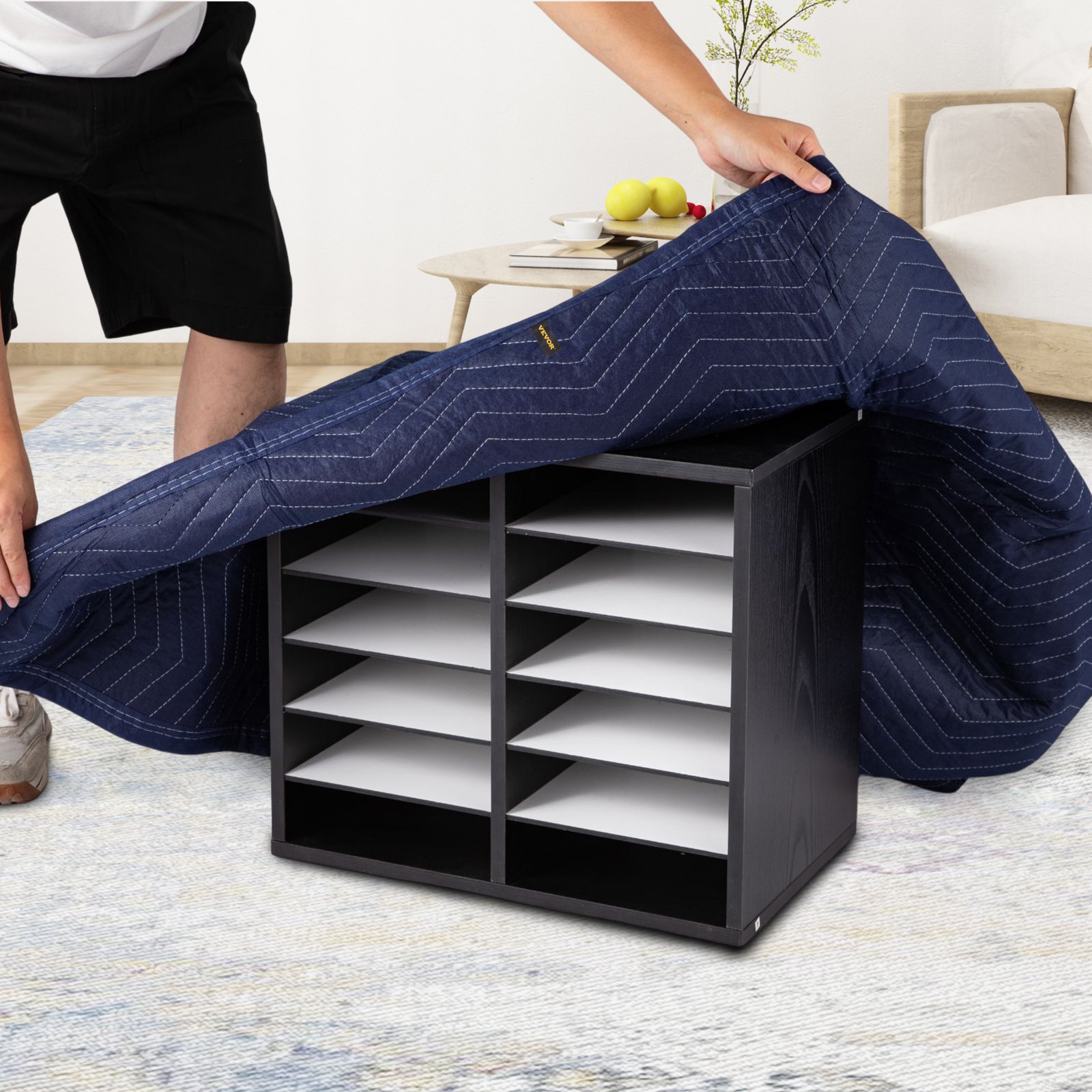 Persona que cubre una estantería negra con una manta de mudanza azul VEVOR en una sala de estar.