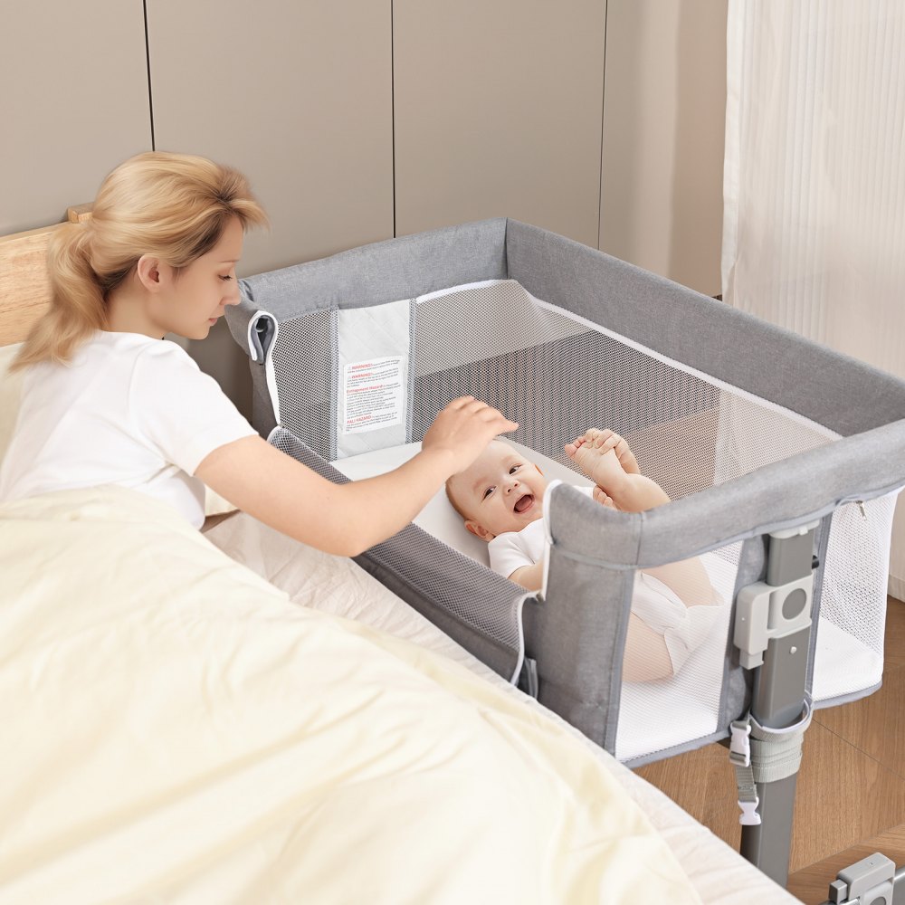 Cuna para bebé VEVOR al lado de la cama con mano de adulto tocando suavemente al bebé dentro de la cuna de malla gris.