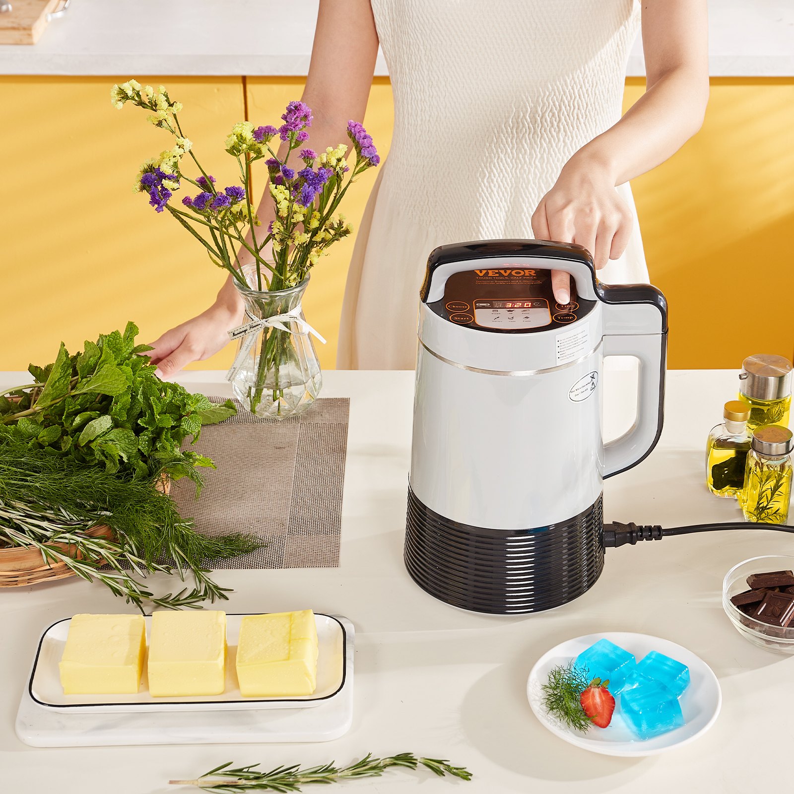 Persona que usa la máquina infusora de mantequilla VEVOR junto a hierbas, mantequilla y gelatina colorida en una cocina.