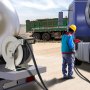 Trabajador reabasteciendo camión utilizando carrete de manguera de combustible VEVOR en un sitio de construcción con un camión volquete al fondo.
