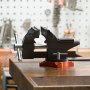 Tornillo de banco VEVOR en un banco de trabajo en un taller, con tablero perforado y herramientas en el fondo borroso.