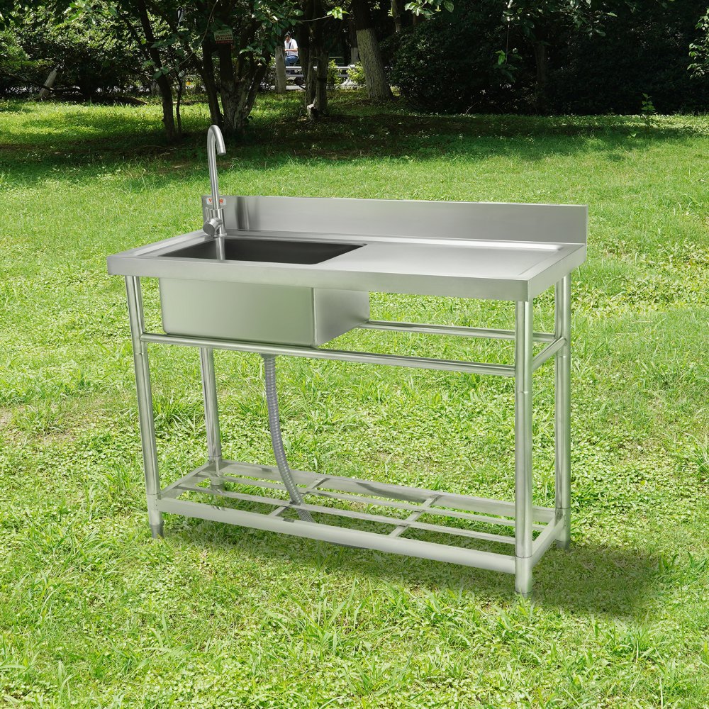 Fregadero de acero inoxidable con grifo, escurridor y estante inferior sobre césped verde en un parque.