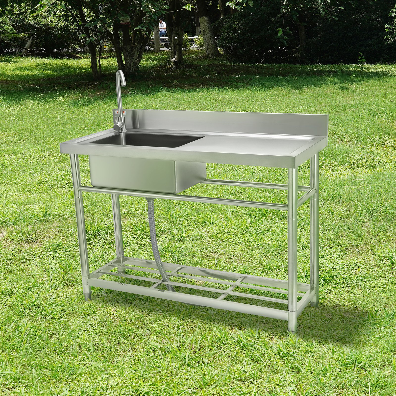 Fregadero de acero inoxidable con grifo, escurridor y estante inferior sobre césped verde en un parque.