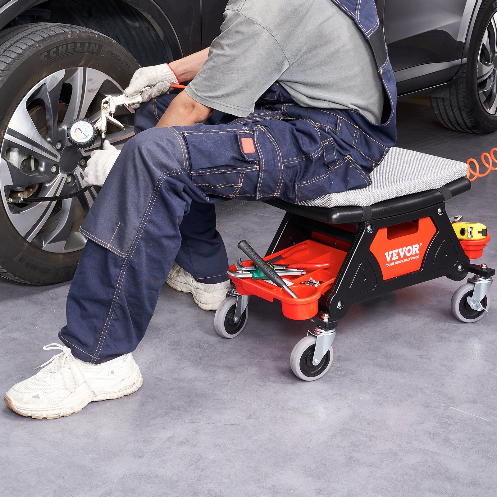 Mecánico usando el taburete de mecánico VEVOR mientras trabaja en un neumático de automóvil, con herramientas almacenadas debajo.