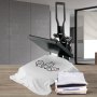 Prensa térmica VEVOR con camiseta impresa y ropa doblada sobre una mesa de madera.