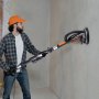 Hombre usando una lijadora de paneles de yeso VEVOR en una pared interior, con gorra naranja, camisa de franela y guantes.