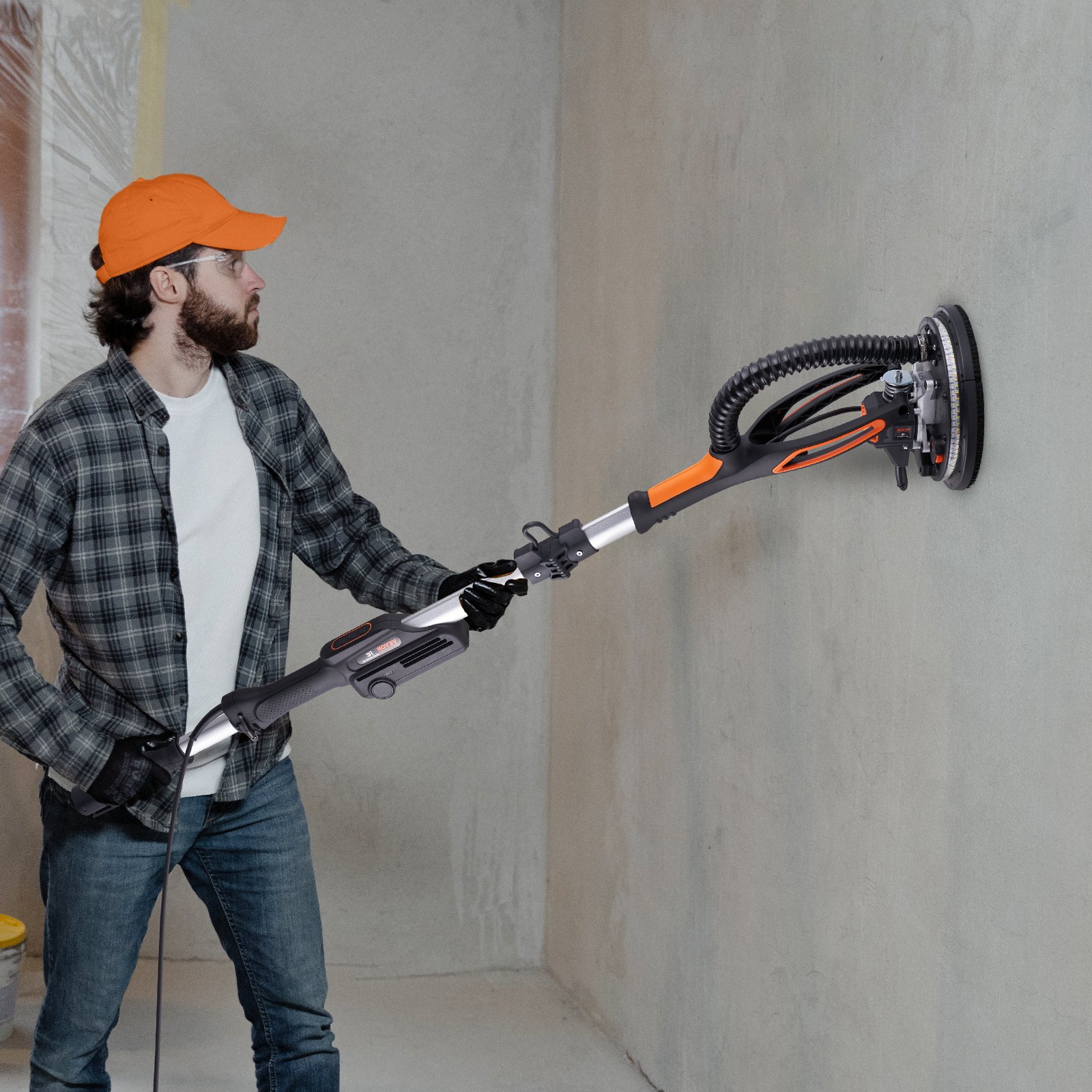 Hombre usando una lijadora de paneles de yeso VEVOR en una pared interior, con gorra naranja, camisa de franela y guantes.