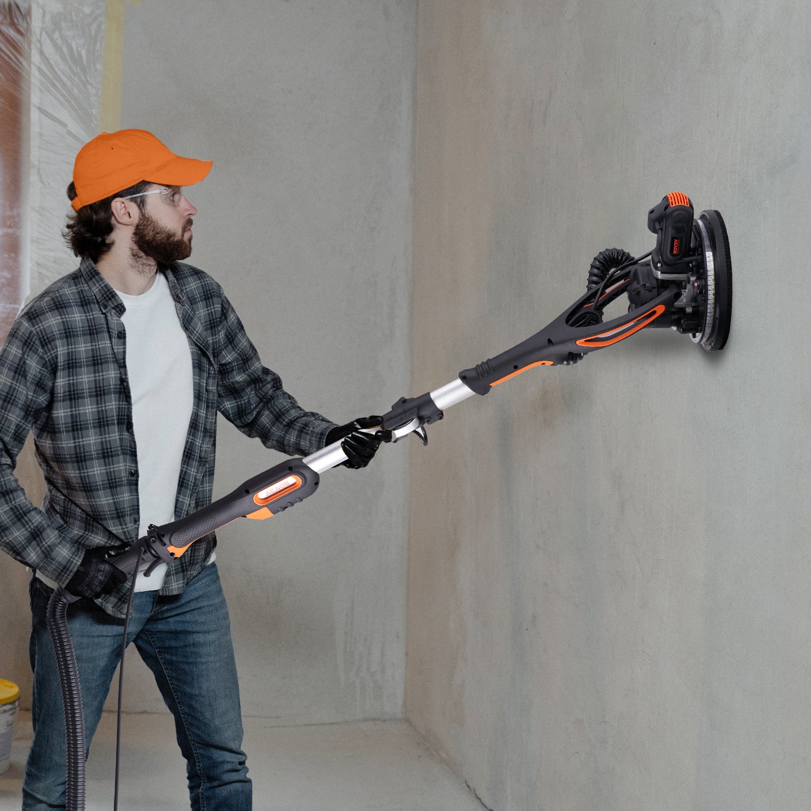 Persona que usa una lijadora de paneles de yeso VEVOR en una pared mientras viste una gorra naranja y una camisa a cuadros.