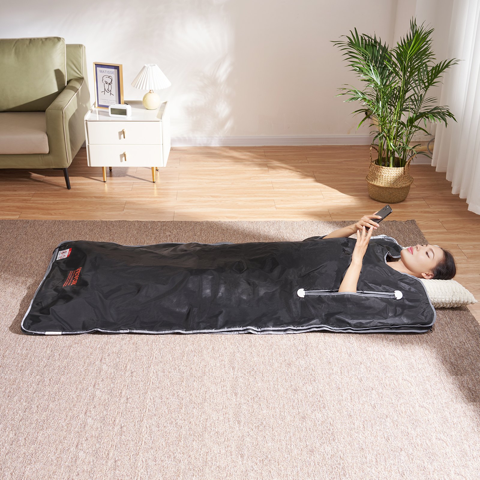 Persona que usa una manta de sauna VEVOR mientras está acostada sobre una alfombra, cerca de un sofá verde, una mesa auxiliar blanca y una planta en maceta.