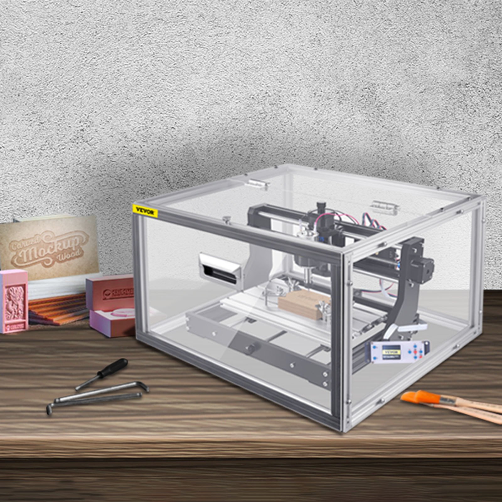 Máquina enrutadora CNC VEVOR en un gabinete transparente sobre una mesa de madera con muestras de tallado.