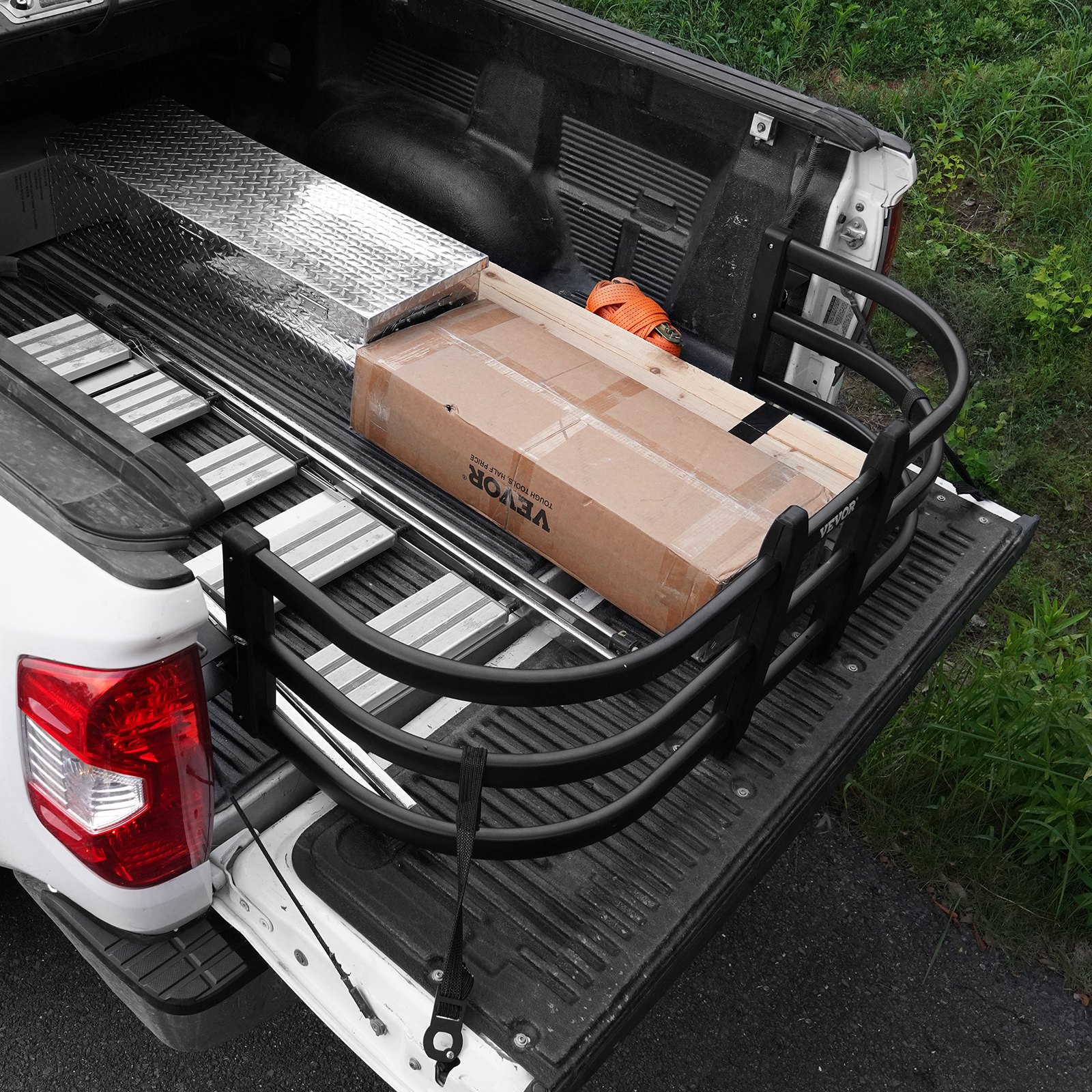 Extensor de caja de camioneta VEVOR que sostiene un artículo en caja, una caja de herramientas de metal y una correa naranja en una camioneta blanca.
