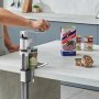 Persona que utiliza un abrelatas comercial VEVOR para abrir una lata en una cocina con diversos productos enlatados.