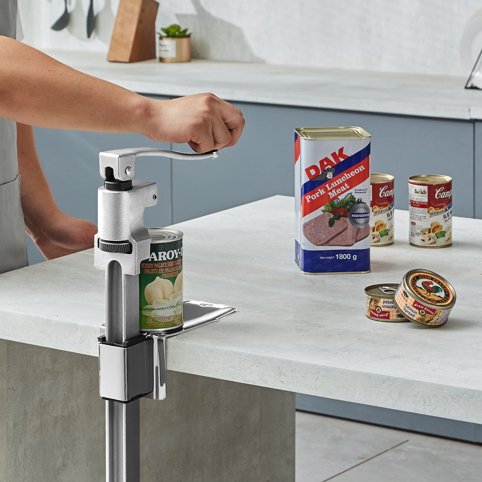 Persona que utiliza un abrelatas comercial VEVOR para abrir una lata en una cocina con diversos productos enlatados.