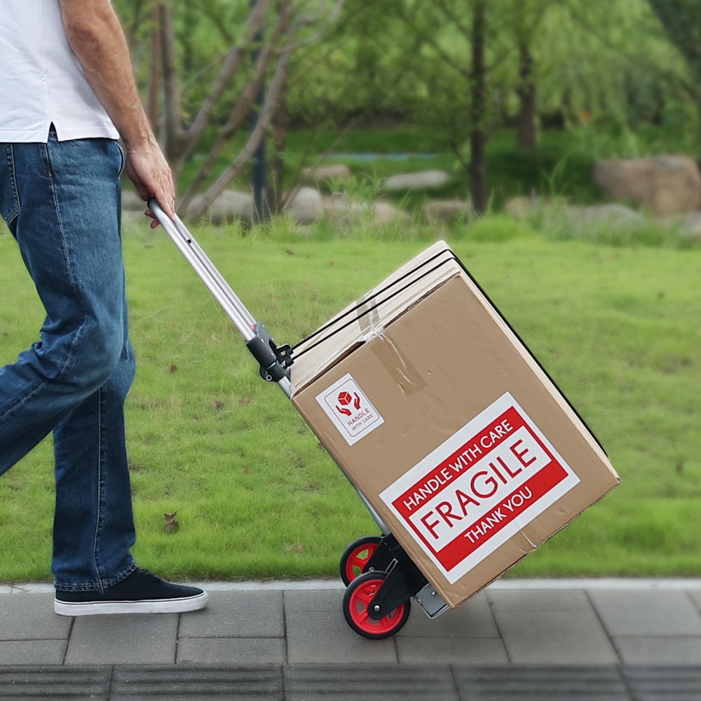 Persona que usa una carretilla manual plegable VEVOR para mover una caja grande y frágil en un camino pavimentado en una zona cubierta de césped.