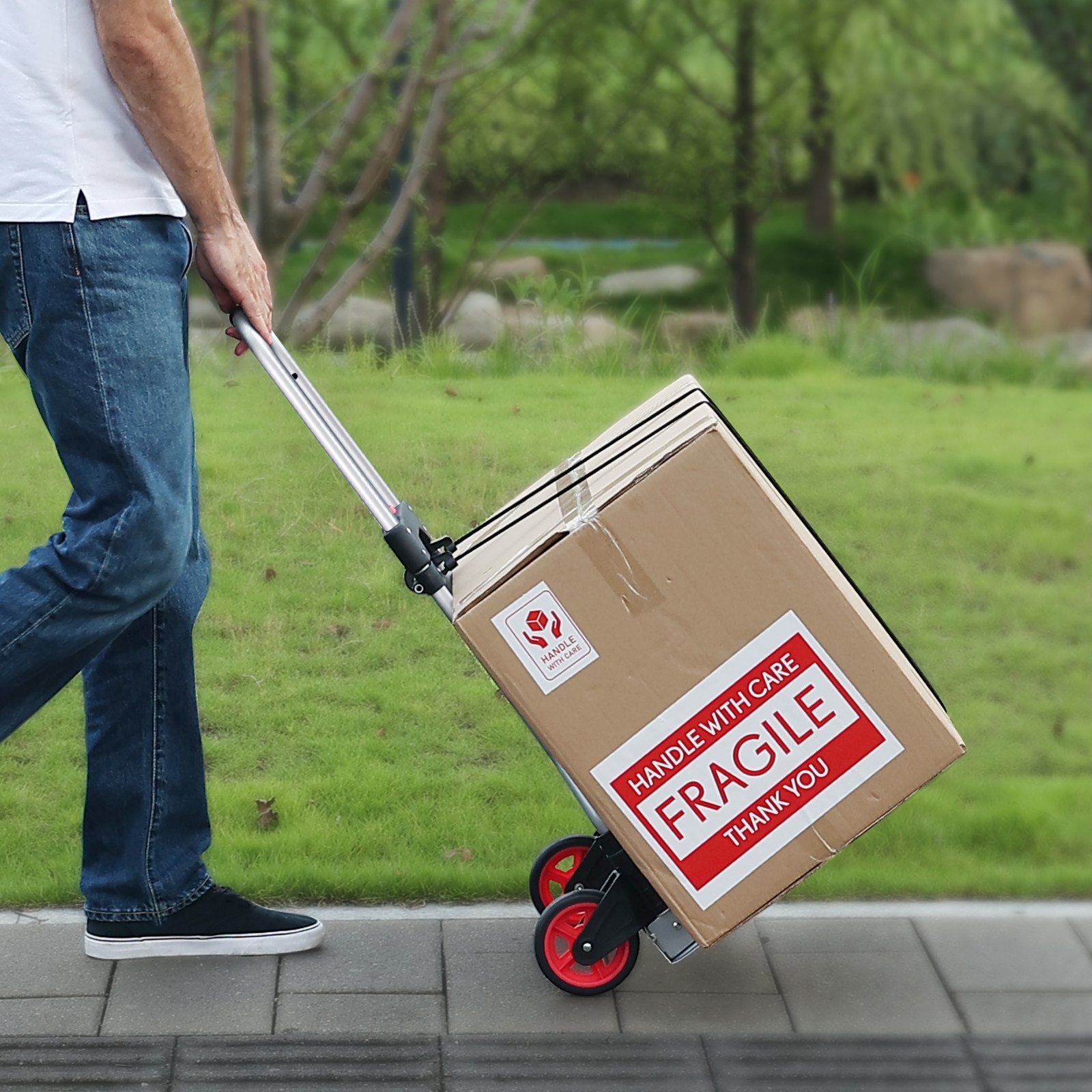 Persona que usa una carretilla manual plegable VEVOR para mover una caja grande y frágil en un camino pavimentado en una zona cubierta de césped.