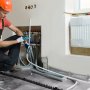Instalador trabajando en un sistema de calefacción con tubería pex VEVOR, conectando líneas debajo de una ventana con un radiador.