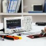 Osciloscopio digital VEVOR que muestra formas de onda junto a herramientas en un escritorio blanco, con estantes en el fondo.