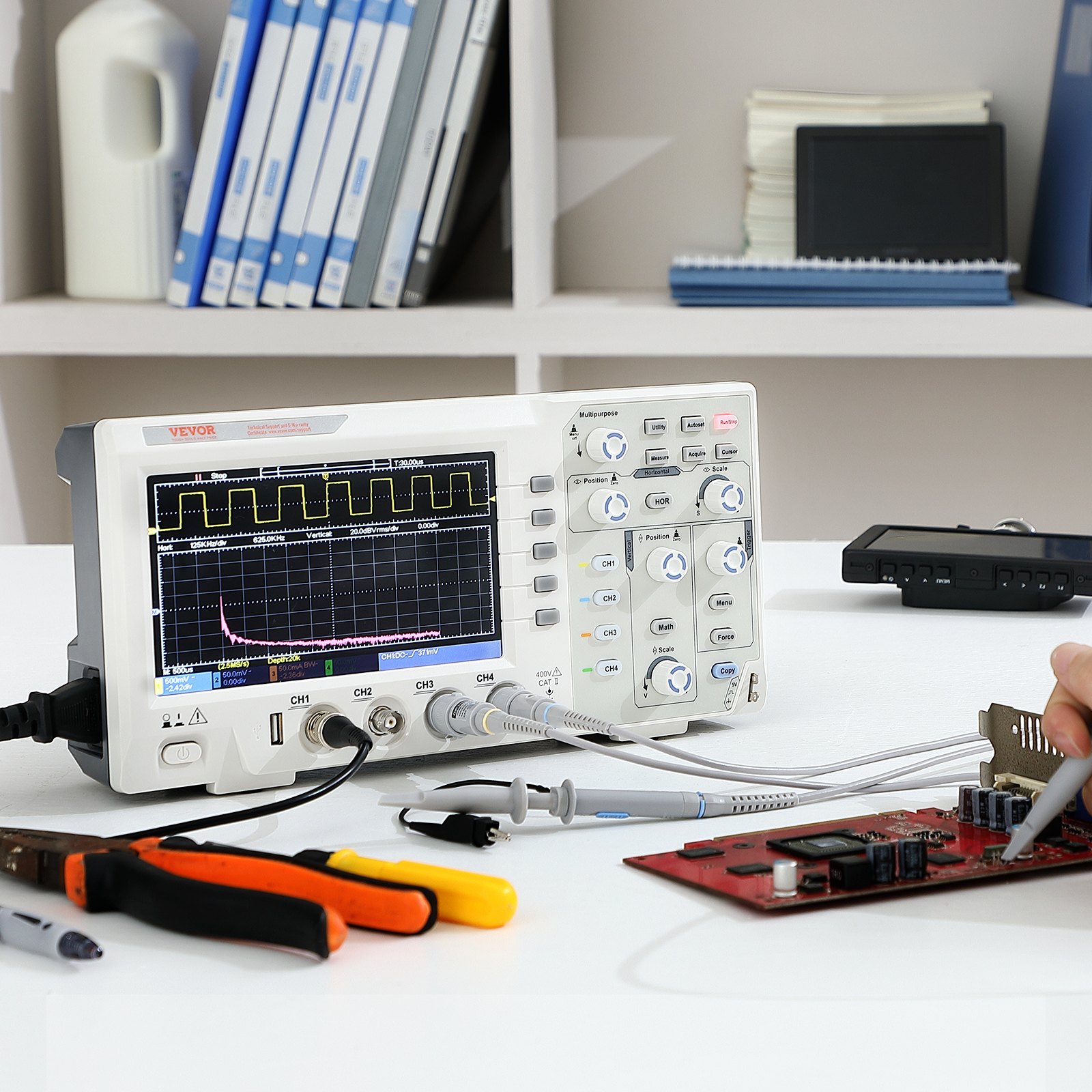 Osciloscopio digital VEVOR que muestra formas de onda junto a herramientas en un escritorio blanco, con estantes en el fondo.