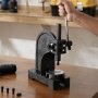 Persona operando una prensa de husillo VEVOR sobre una mesa de madera en un taller, con herramientas cerca.