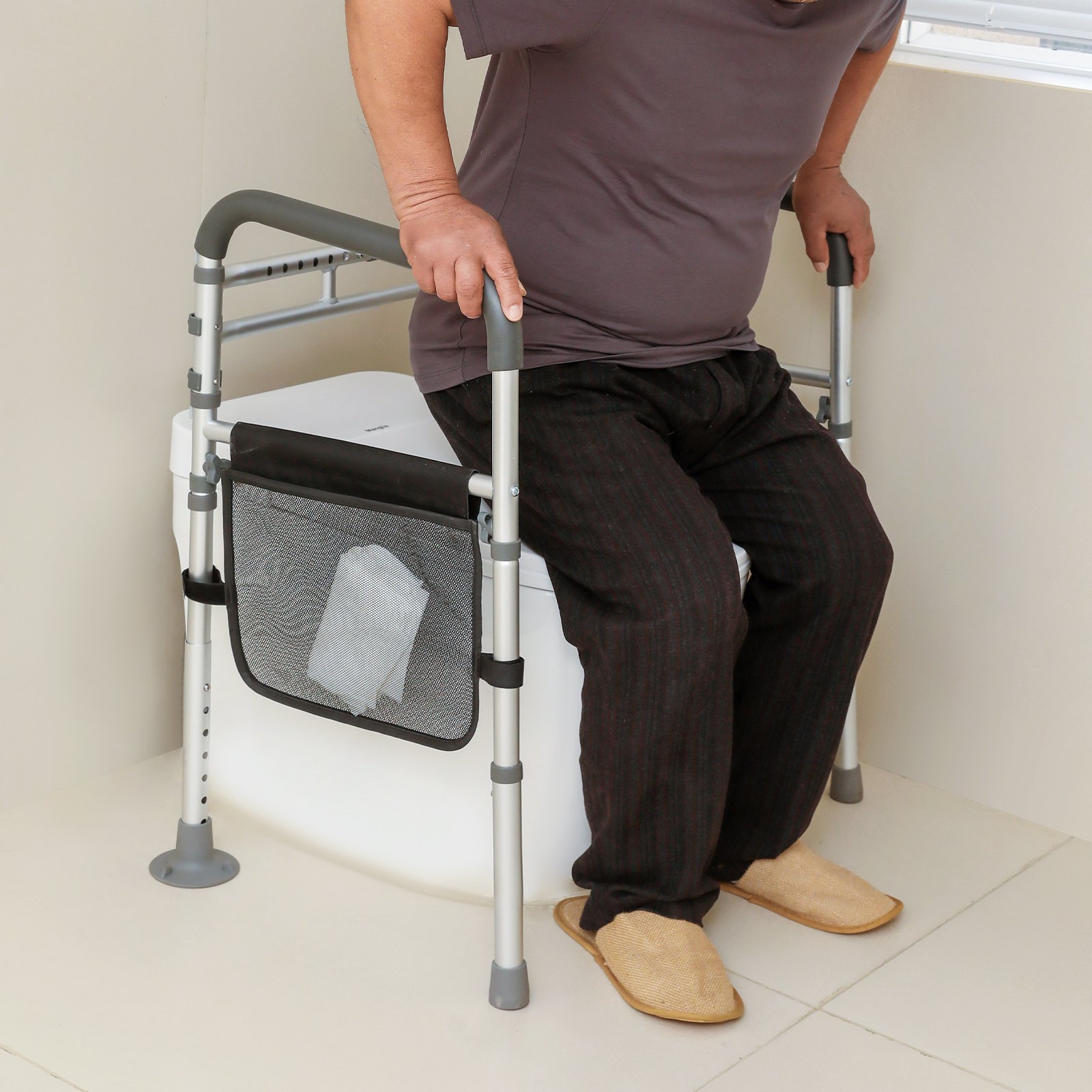 Persona mayor que utiliza el pasamanos de seguridad para inodoro VEVOR para recibir ayuda, con un organizador de malla que sostiene el pañuelo, en un entorno de baño.