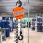 Polipasto de cadena manual VEVOR en un almacén industrial con barriles y paletas al fondo.