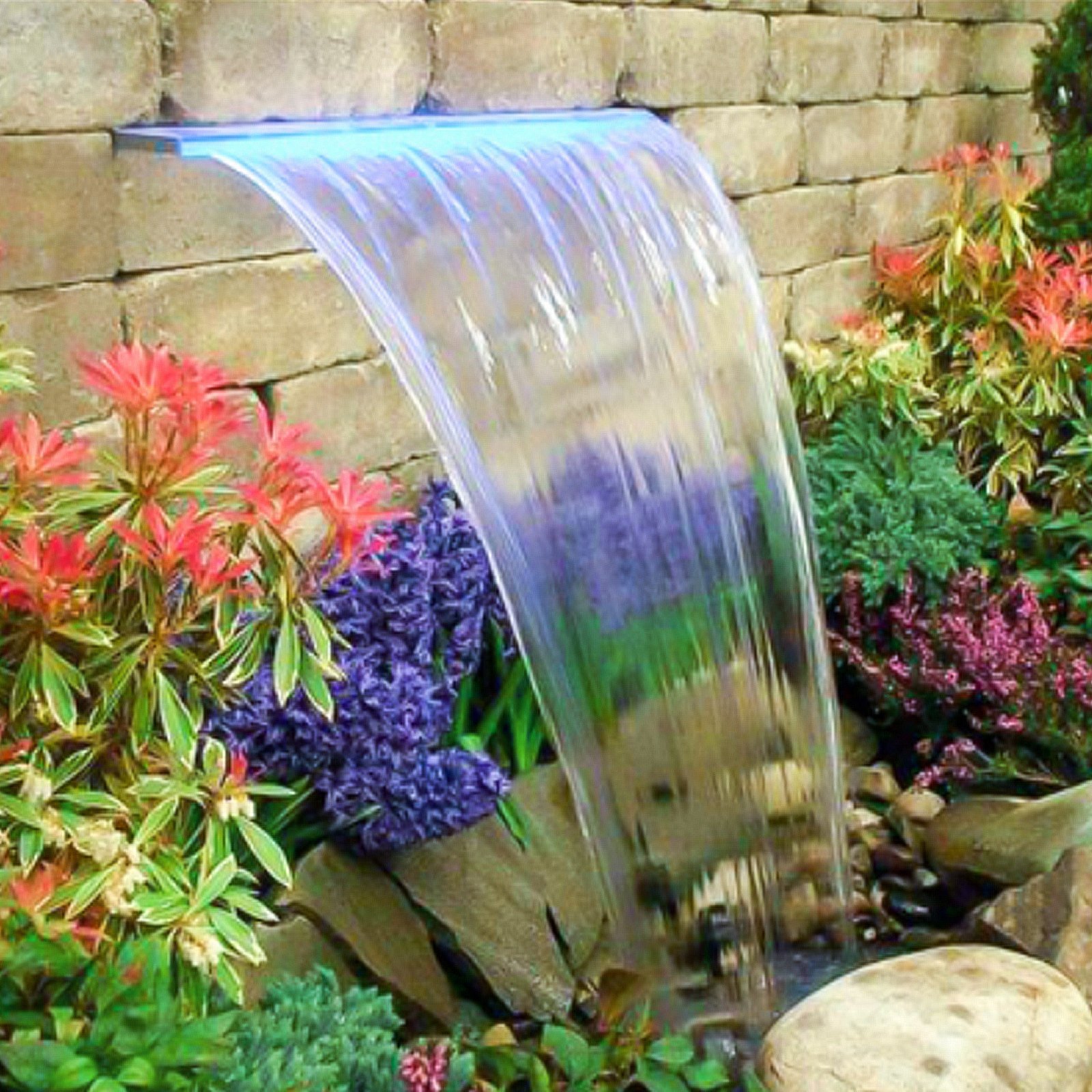 Aliviadero de fuente VEVOR con agua en cascada en medio de coloridas flores de jardín y un fondo de piedra.