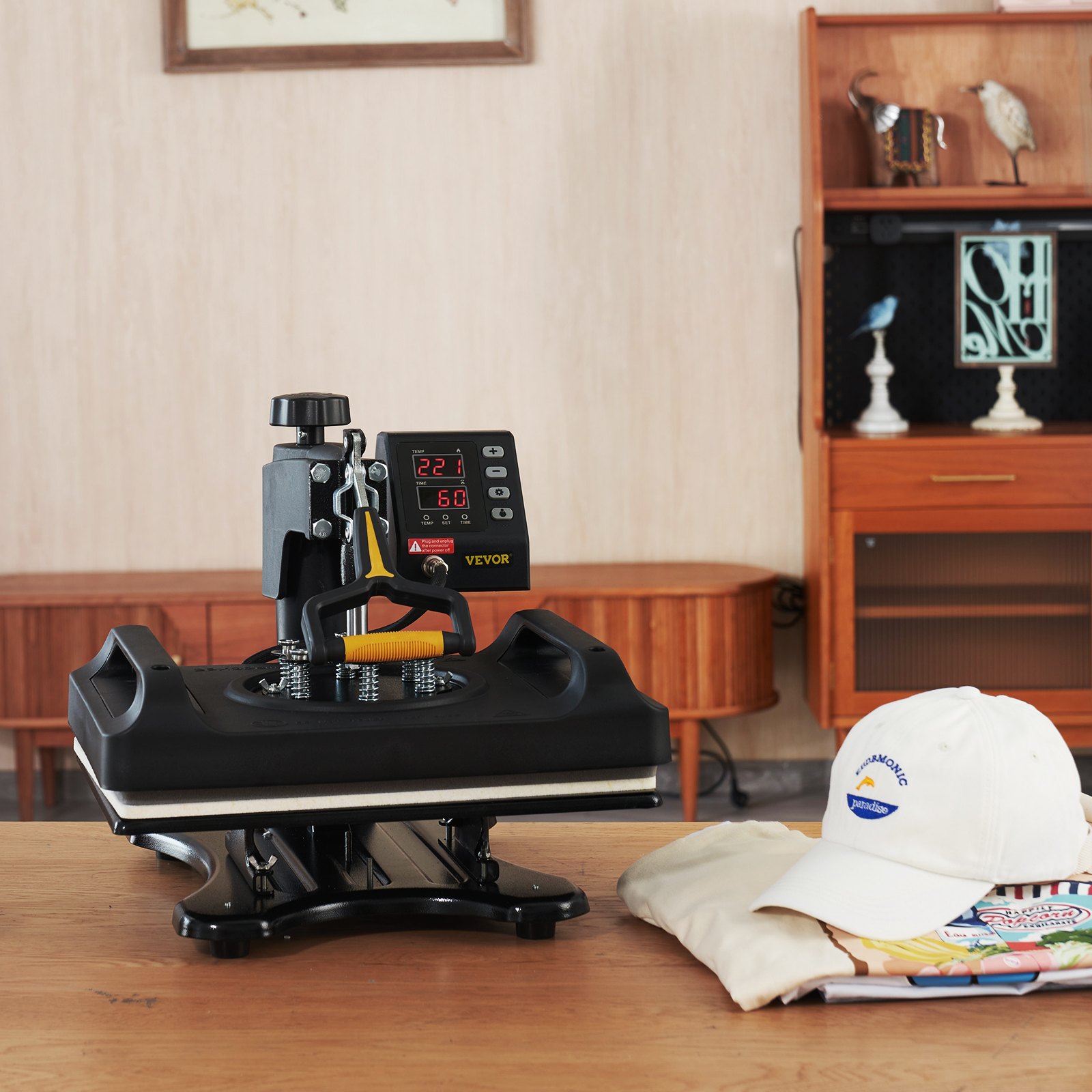 Máquina de prensado térmico VEVOR sobre una mesa de madera con una gorra y una camiseta cerca.