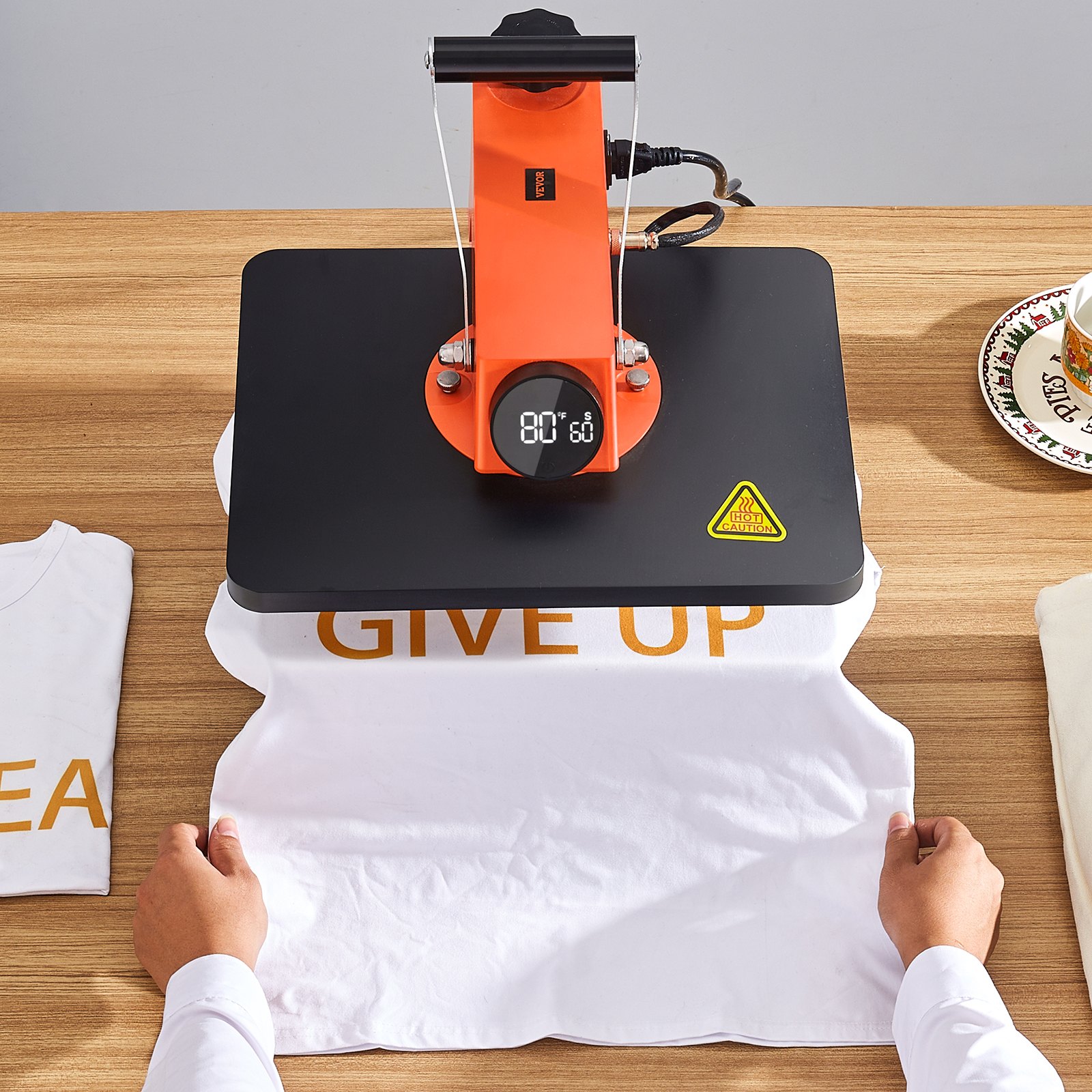 Máquina de prensado térmico VEVOR sobre mesa de madera con pantalla digital, prensando una camiseta blanca con texto.