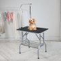 Perro pequeño sentado en una mesa de aseo para mascotas VEVOR con brazo ajustable, cerca de toallas de colores en perchas.