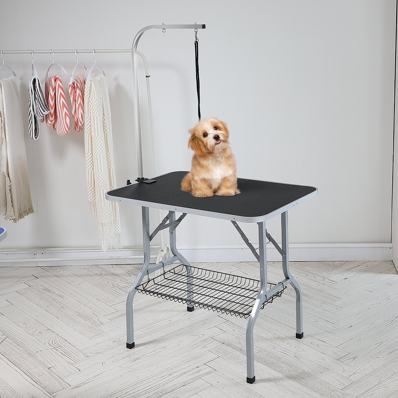 Perro pequeño sentado en una mesa de aseo para mascotas VEVOR con brazo ajustable, cerca de toallas de colores en perchas.