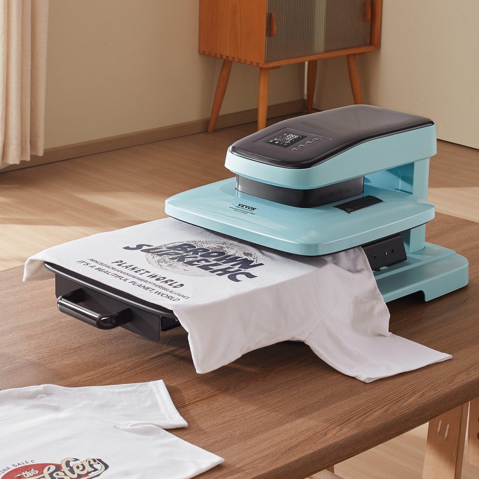 Máquina de prensado térmico automática VEVOR sobre una mesa de madera imprimiendo una camiseta blanca con texto en una habitación llena de luz.