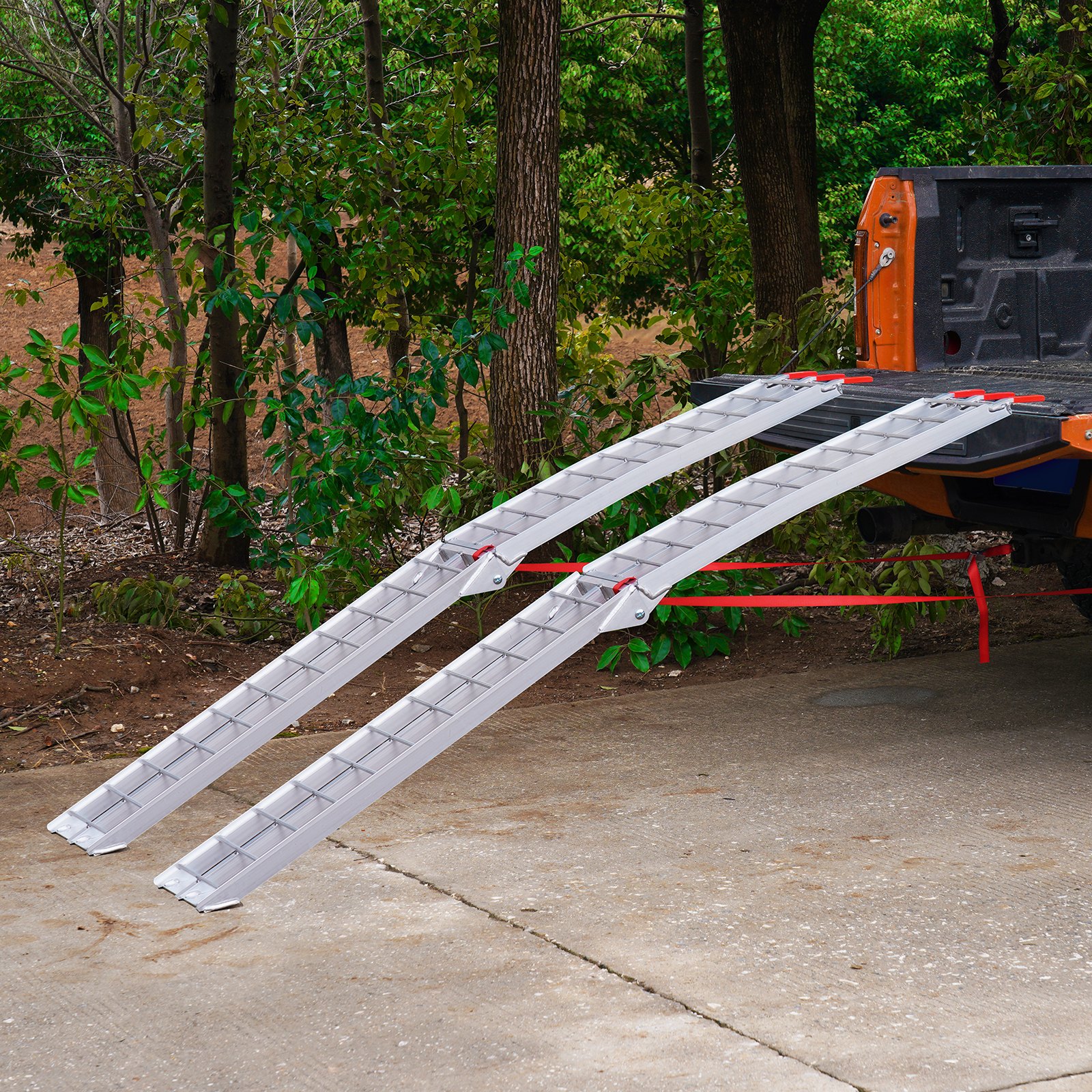 Rampas de carga de aluminio VEVOR unidas a un camión naranja, que se extienden hasta el hormigón con árboles al fondo.