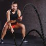 Vrouw oefent met VEVOR battle rope in de sportschool, draagt een zwart topje en oranje korte broek, tegen een bakstenen muur.