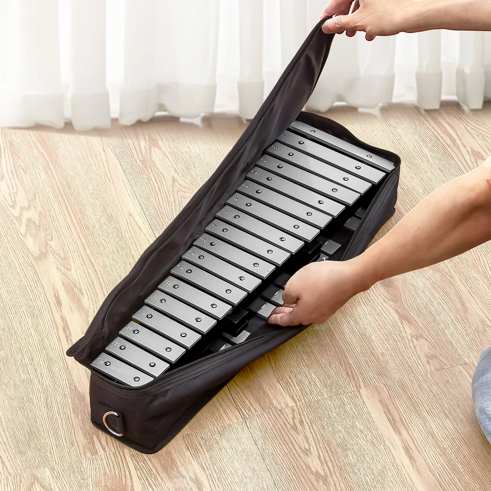 hands placing a VEVOR glockenspiel xylophone into a black carrying case on a wooden floor.