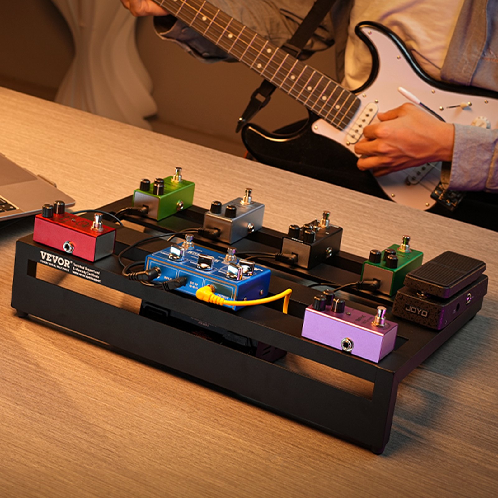 VEVOR guitar pedal board with colorful effects pedals on a table, guitarist playing a black electric guitar.