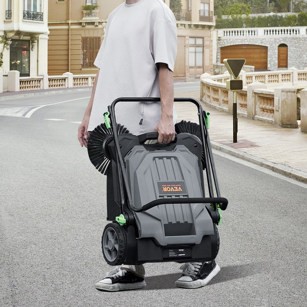person holding a VEVOR floor sweeper on an empty street with residential buildings in the background.