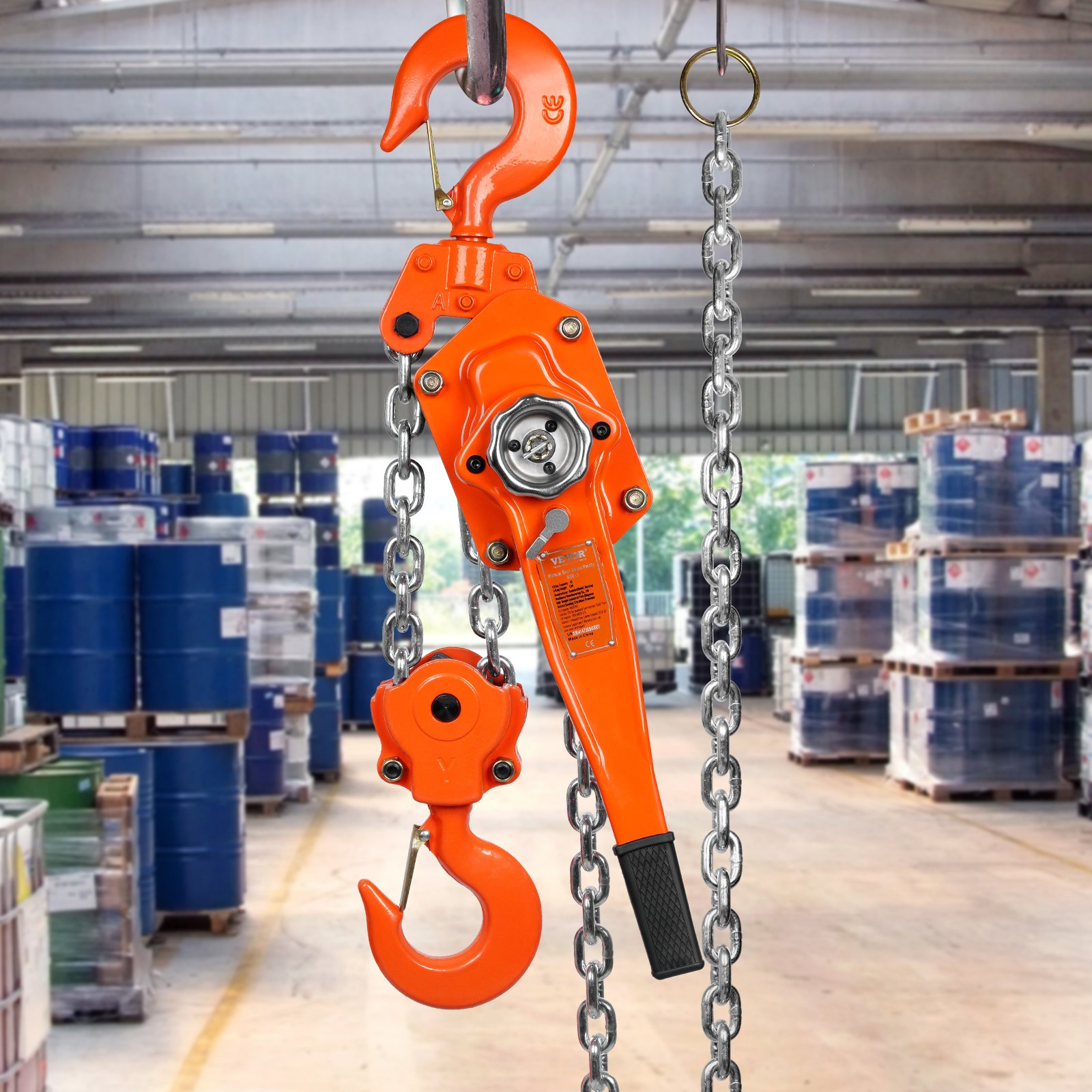 orange VEVOR chain hoist in a warehouse with stacked blue barrels in the background.