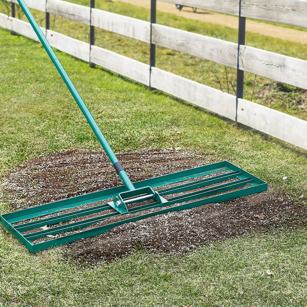 VEVOR lawn leveling rake used to spread soil over a grass lawn, next to a wooden fence.