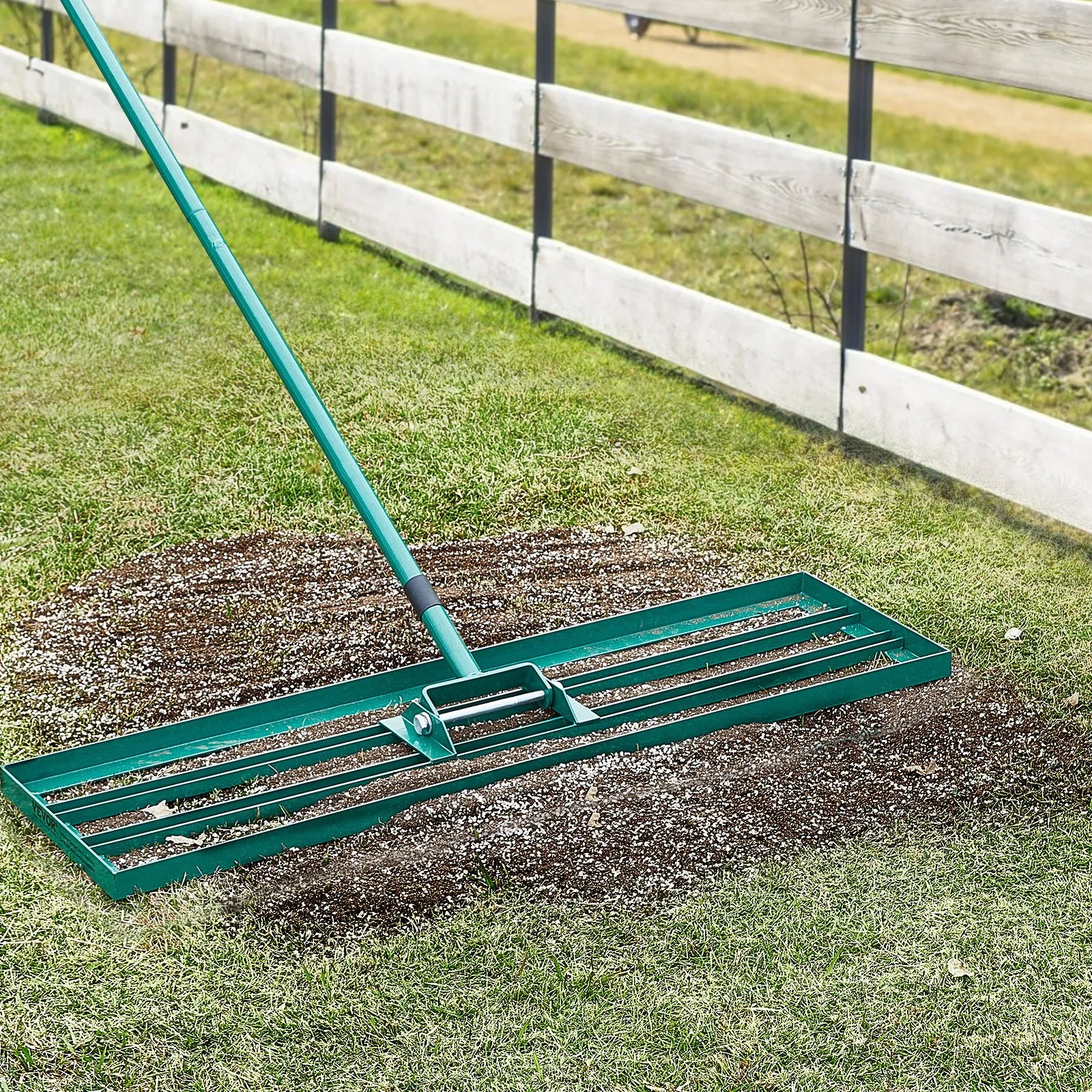 VEVOR lawn leveling rake used to spread soil over a grass lawn, next to a wooden fence.