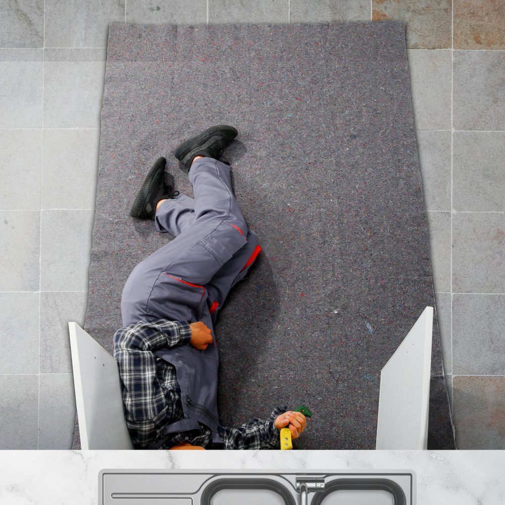 worker lying on VEVOR moving blankets, wearing uniform and holding a tool, tiles in the background.
