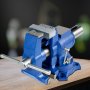 blue VEVOR bench vise mounted on a wooden surface with a dark blurred background.