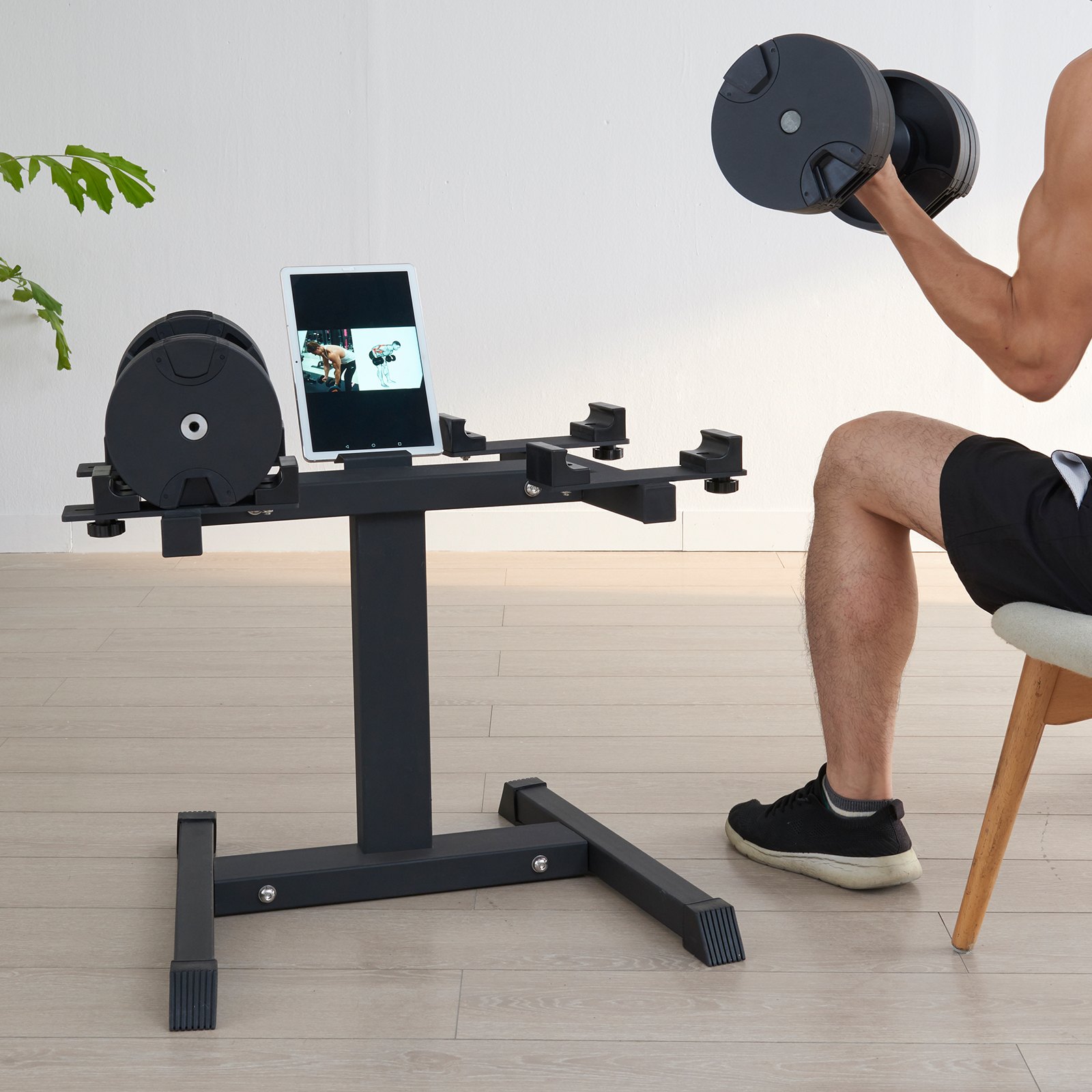 man exercising with dumbbells next to a VEVOR dumbbell stand holding a tablet in a bright room.