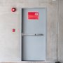 gray door with a VEVOR panic exit device, red exit sign above, and fire alarm equipment nearby.