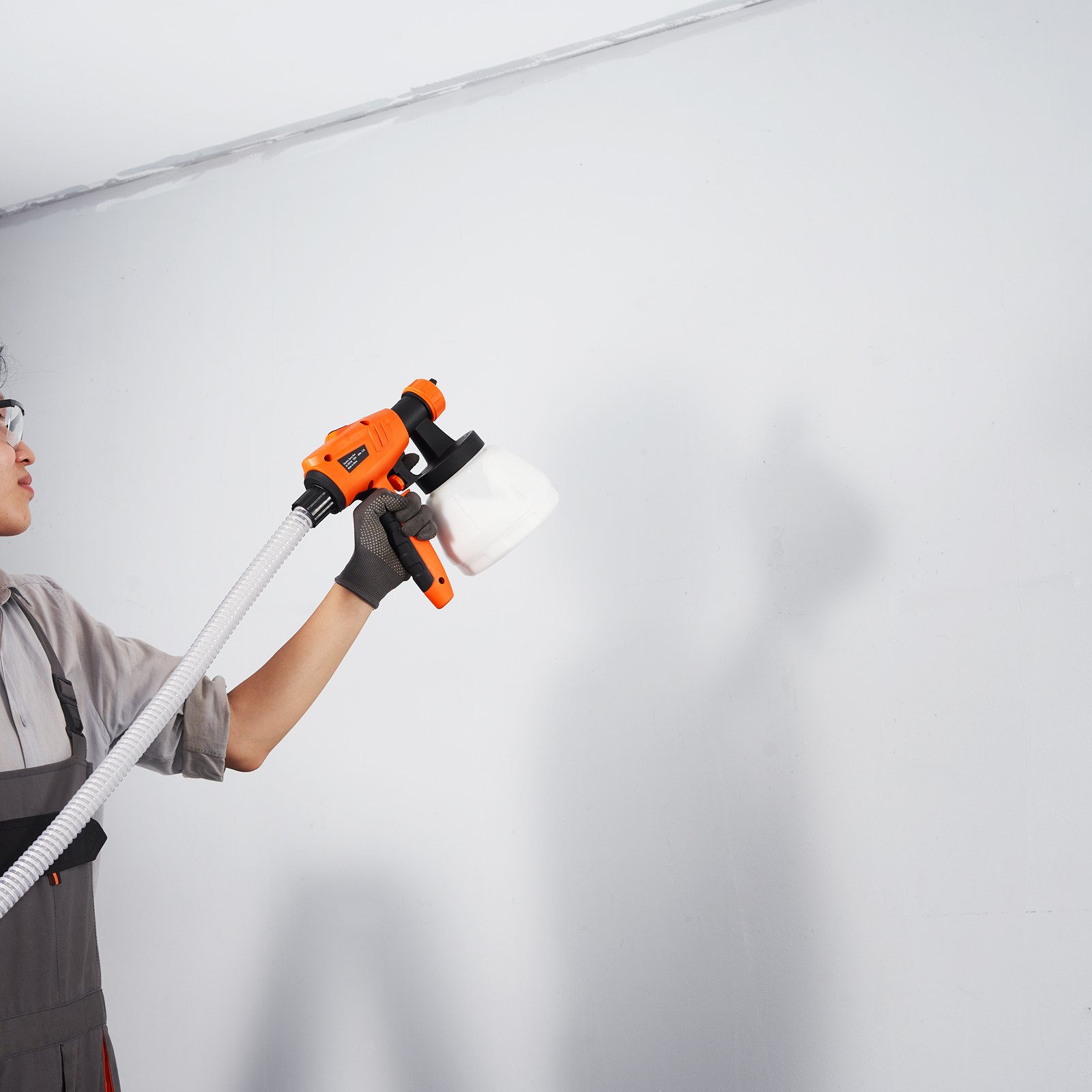Persona que utiliza un pulverizador de pintura VEVOR, con guantes, pintando una pared blanca.
