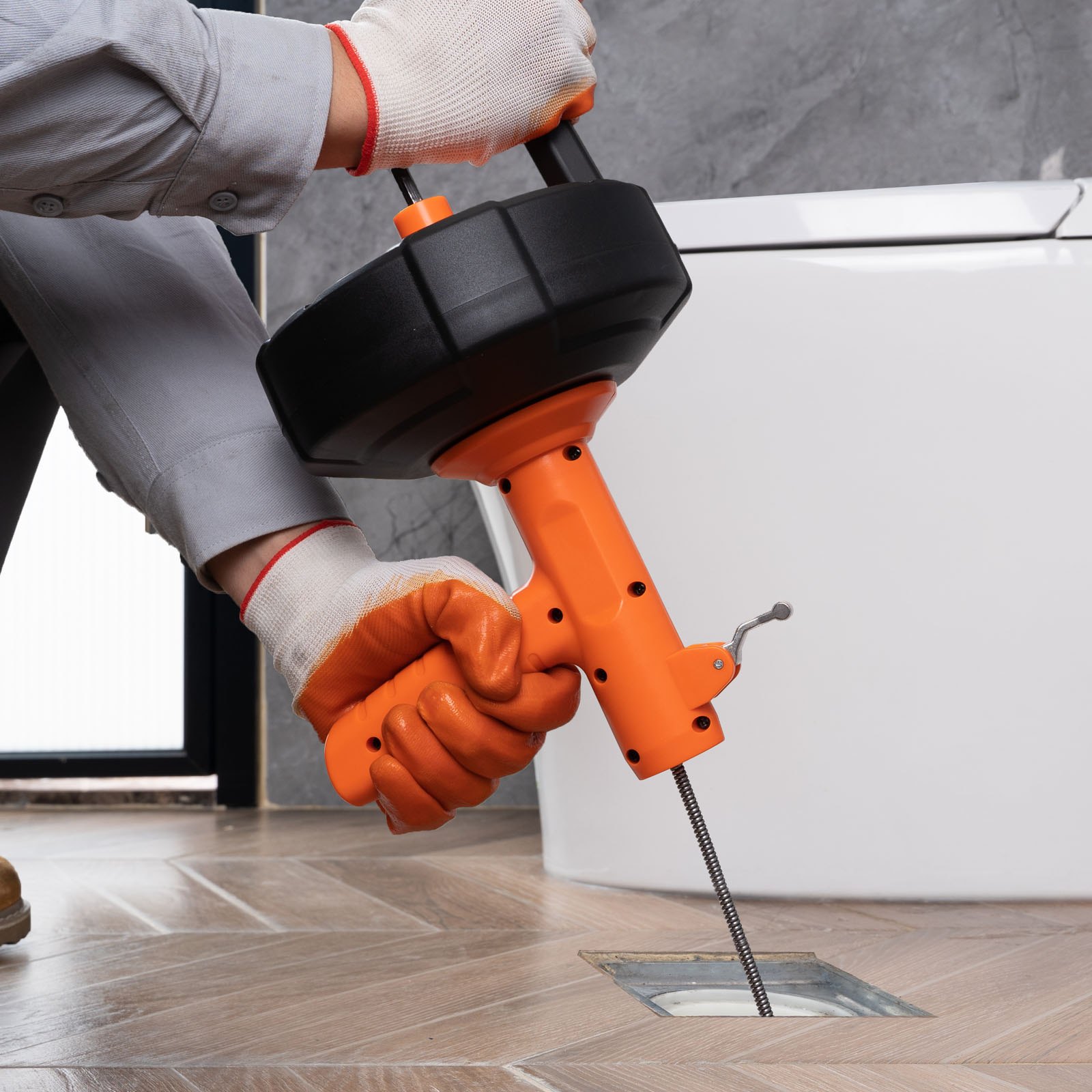 person using VEVOR drain auger to unclog a floor drain near a toilet.