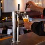 hand pouring beer from VEVOR dual taps draft beer tower on a countertop in a dimly lit bar.