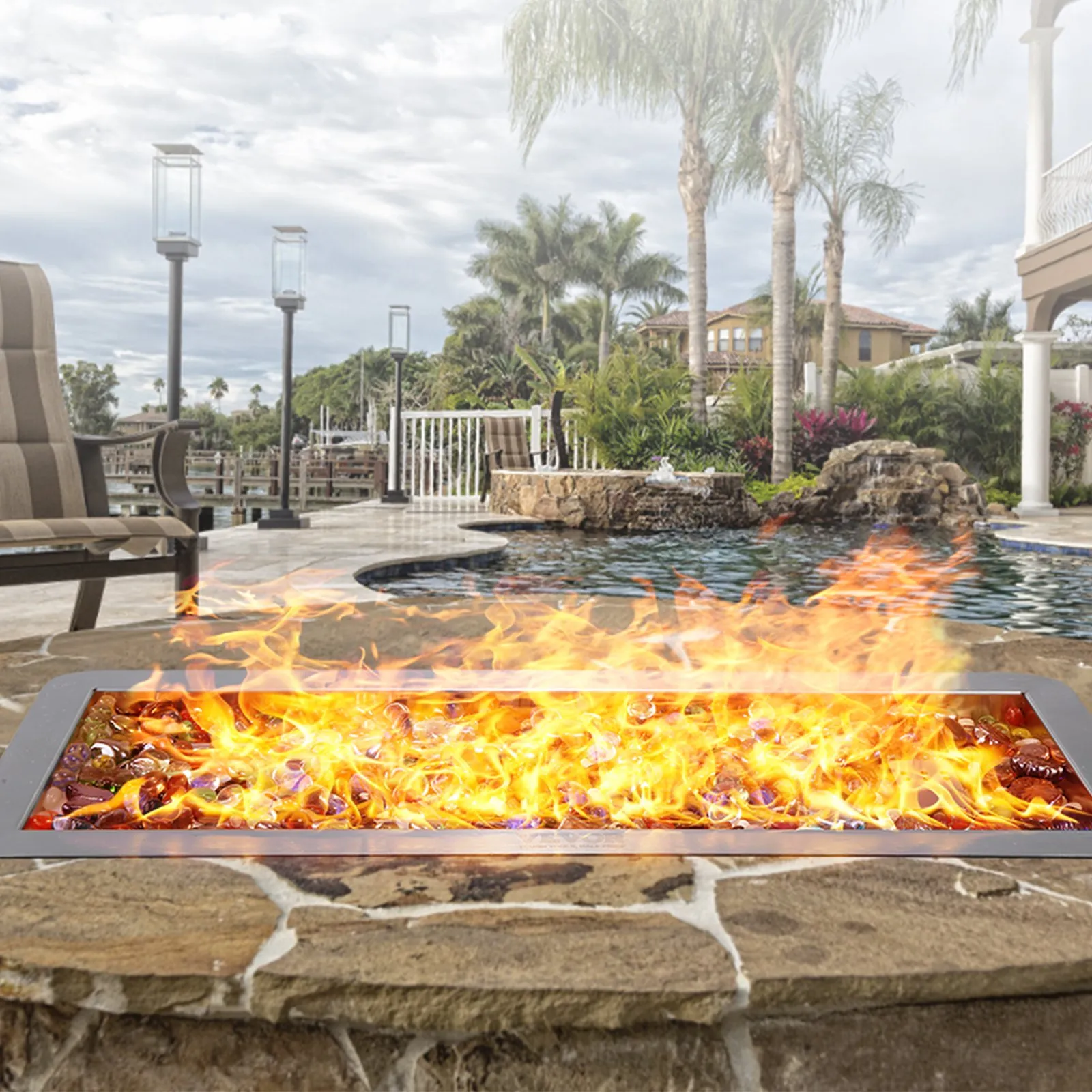 VEVOR fire pit pan with vibrant flames by a luxurious poolside, surrounded by palm trees and seating.