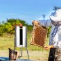 beekeeper using VEVOR honey extractor, holding honeycomb frame near a smoker in an open field.
