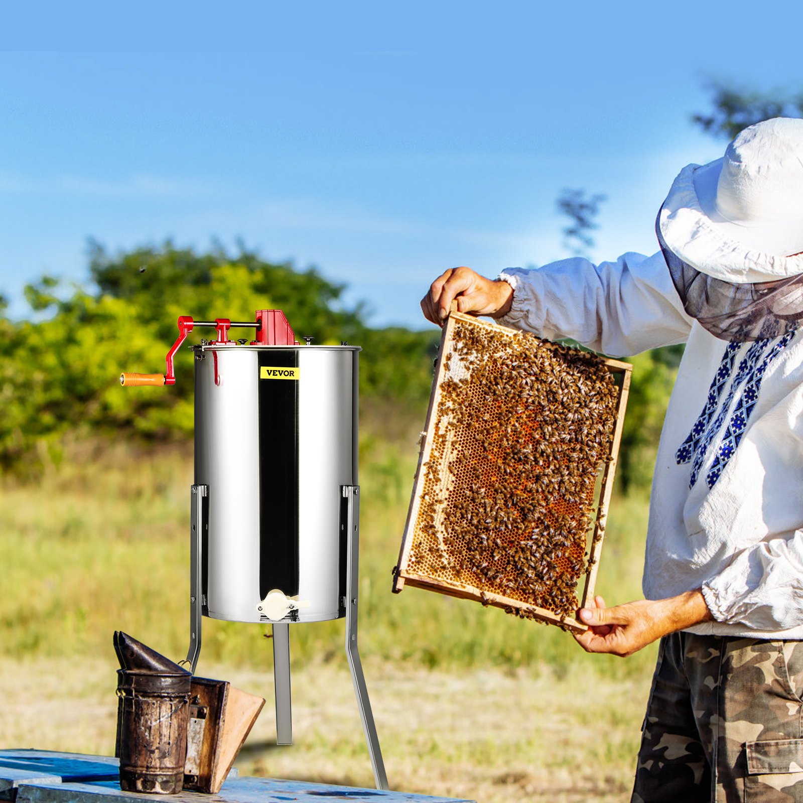 beekeeper using VEVOR honey extractor, holding honeycomb frame near a smoker in an open field.